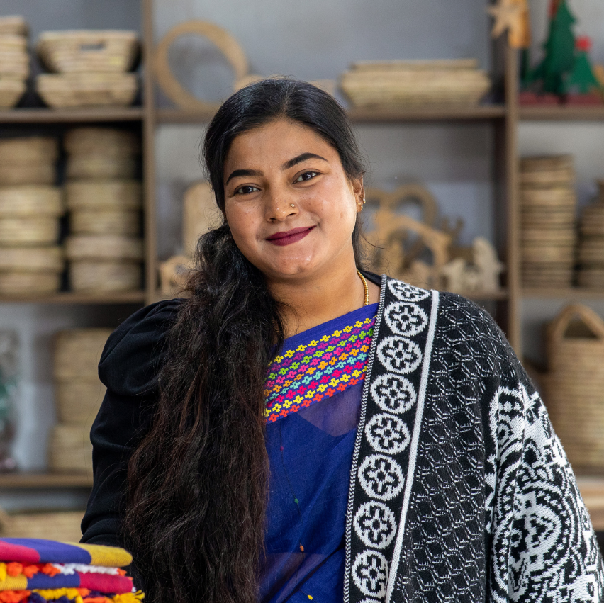 Woman in traditional clothing standing in an Others Trade for Hope shop in Bangladesh with shelves of woven items.